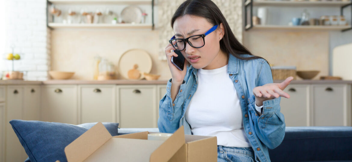 Unsatisfied woman received a broken parcel delivered from an online online store, Asian quarrels with the customer support service, talks to the operator on the phone, sitting home in the kitchen